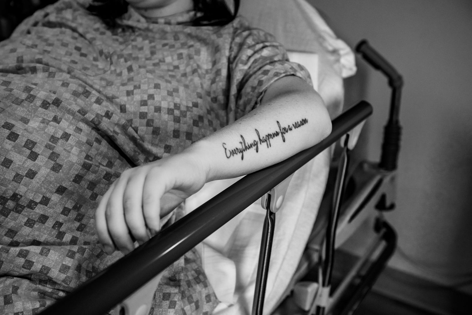 Black and white photo showing hospital patient's arm with text tattoo resting on bed rail in medical gown setting.