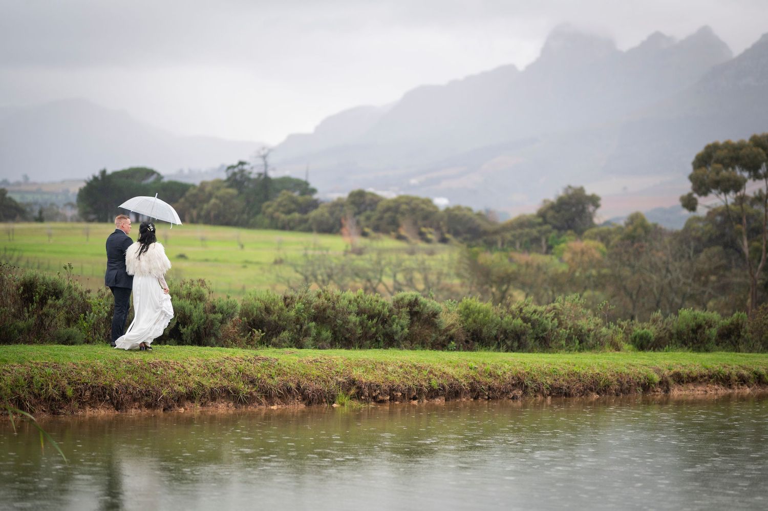 Two people with a white umbrella stand along a scenic riverbank with misty mountains in the background.