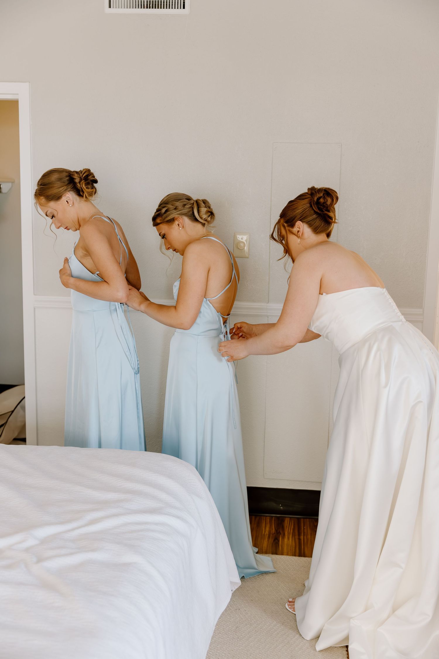 Bridesmaids in light blue dresses help zip up and adjust gowns while getting ready in a sunlit bedroom.