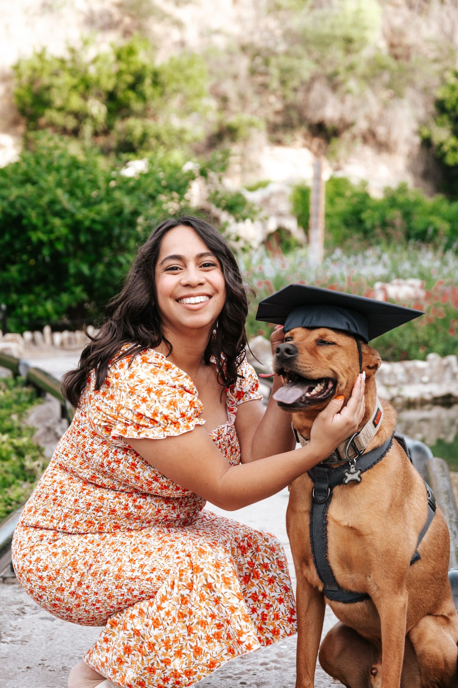 A person in a floral dress celebrates with their brown dog wearing a graduation cap outdoors.