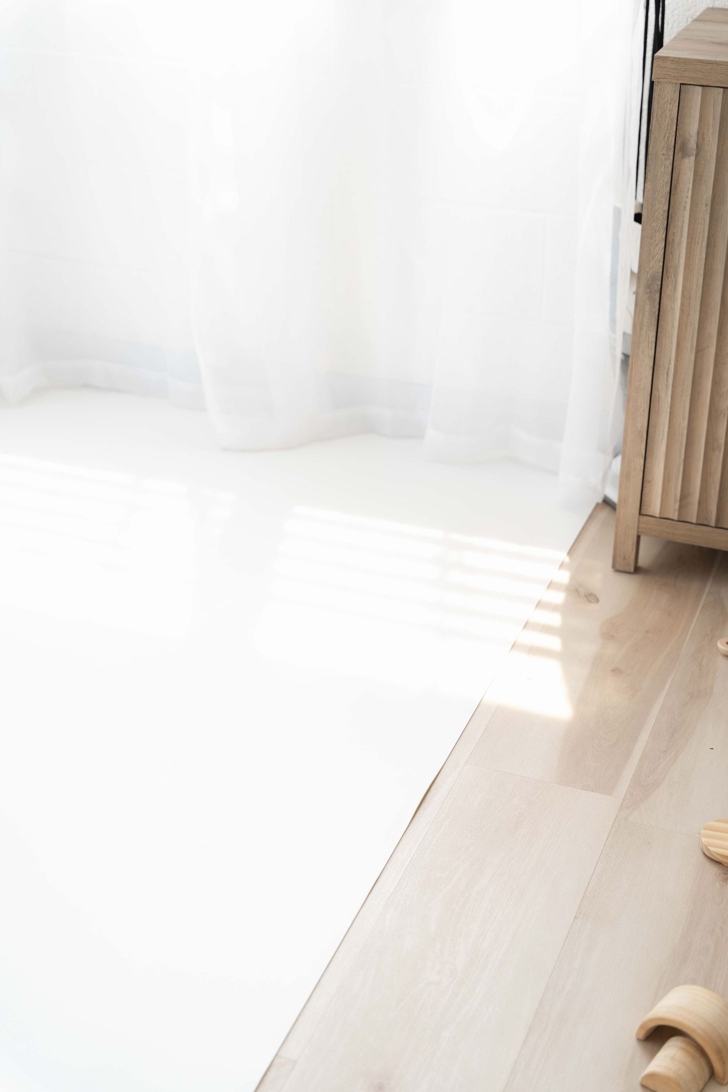 Sunlight streams through white sheer curtains onto a light wood floor beside a wooden cabinet.