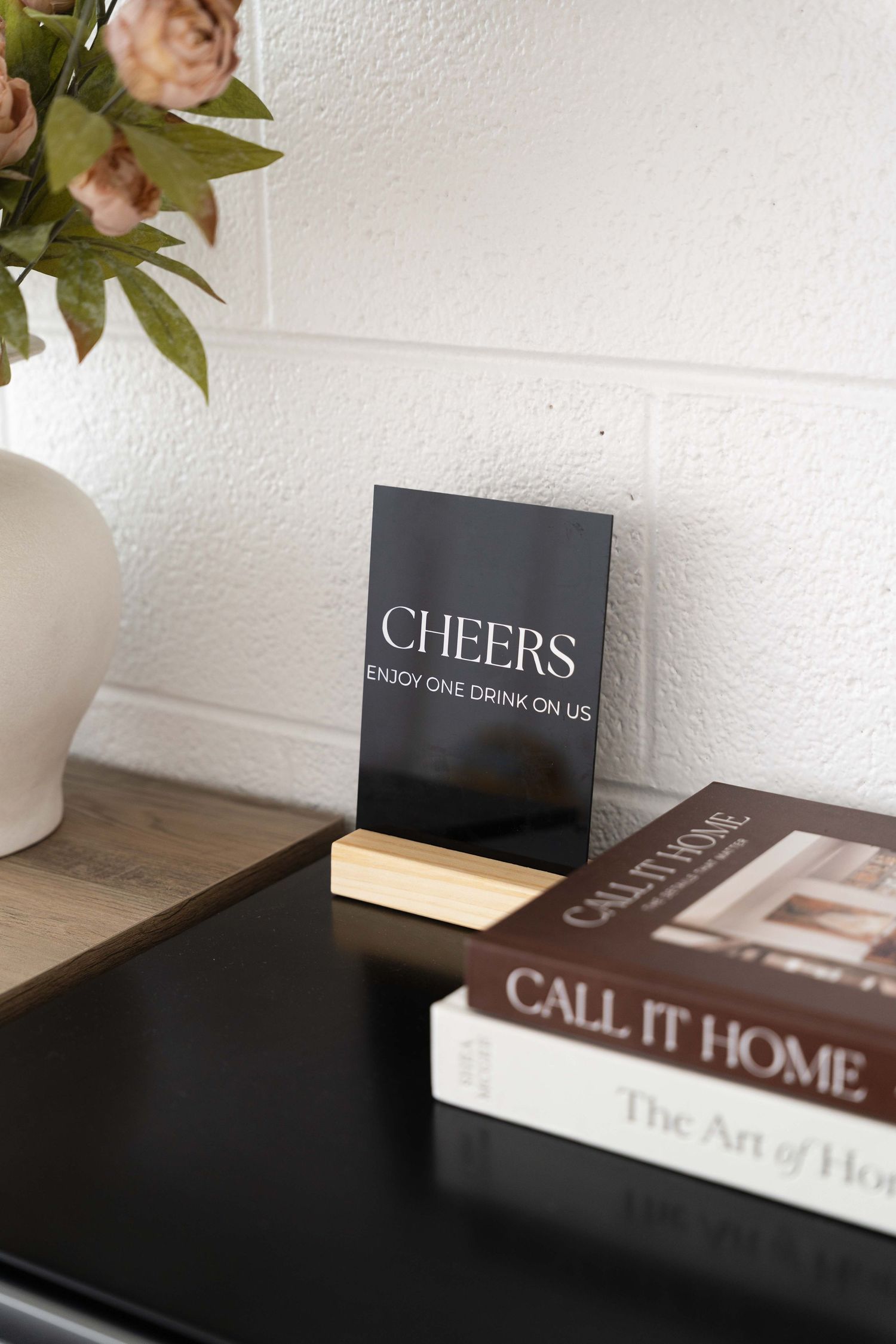 A black 'Cheers' sign displayed alongside books on a wooden surface with pink florals in a white vase.