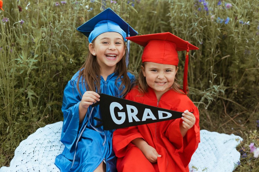 Two graduates in red and blue caps and gowns hold GRAD letters while sitting and smiling on a white blanket outdoors.