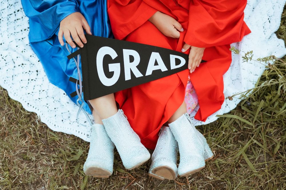 Graduates in contrasting red and blue gowns share a playful moment sitting on grass together.