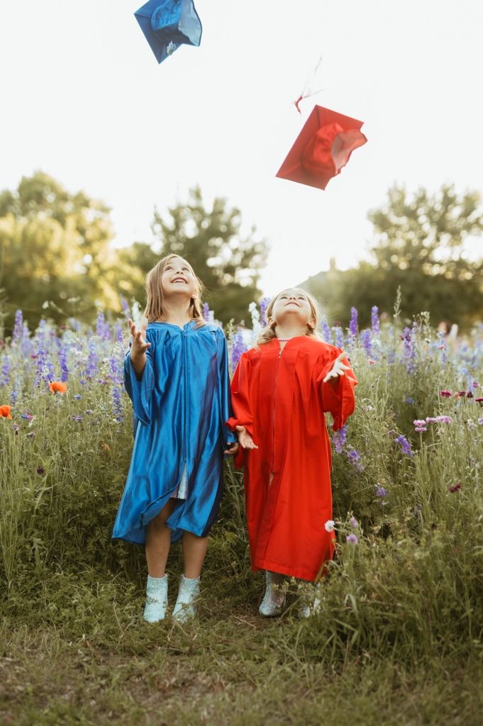 Two graduates in blue and red gowns celebrate in a wildflower field at sunset as their caps soar through the air.