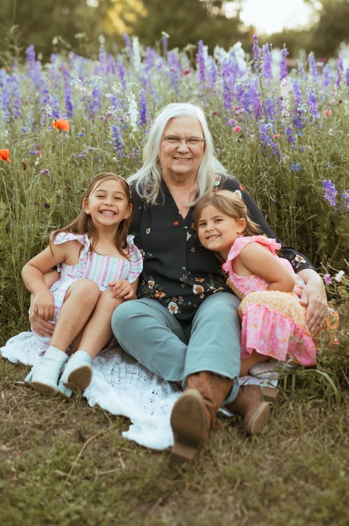 Three generations share a joyful moment sitting together among blooming purple wildflowers on a summer evening.