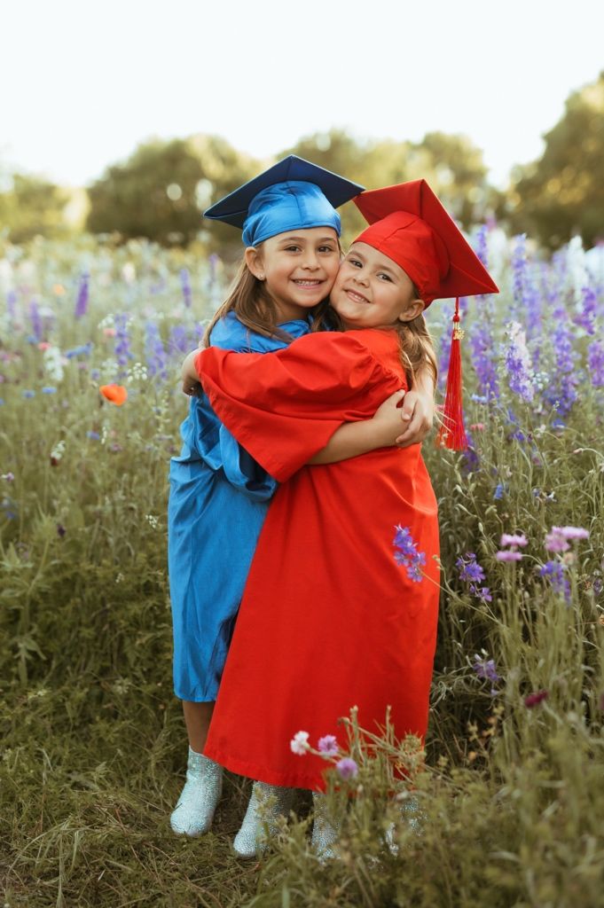Two graduates in red and blue caps and gowns hug while standing in a field of purple wildflowers.