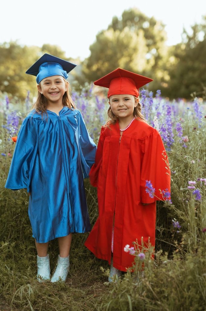 Two young graduates in blue and red caps and gowns stand together in a field of purple wildflowers at sunset.