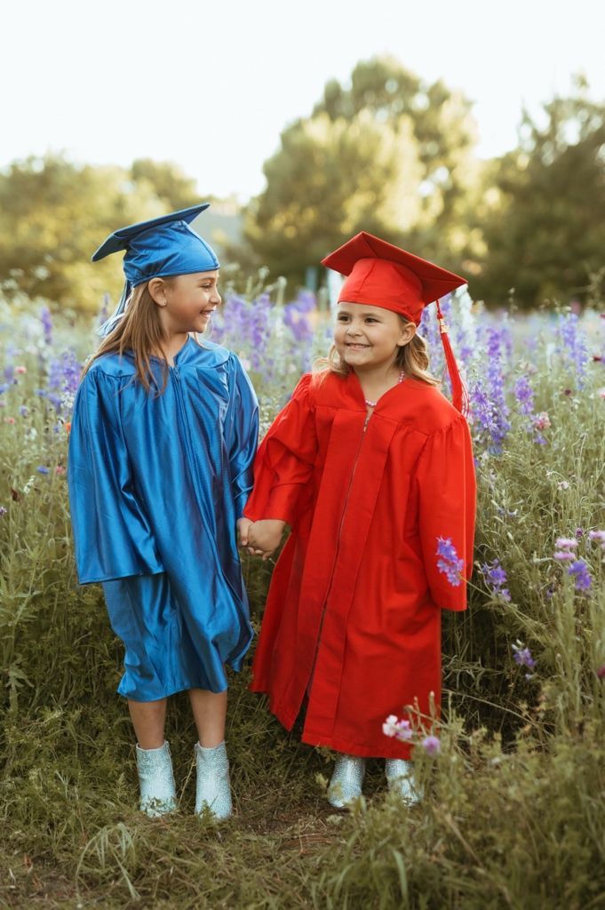 Two graduates in blue and red caps and gowns stand together in a field of purple wildflowers at sunset.