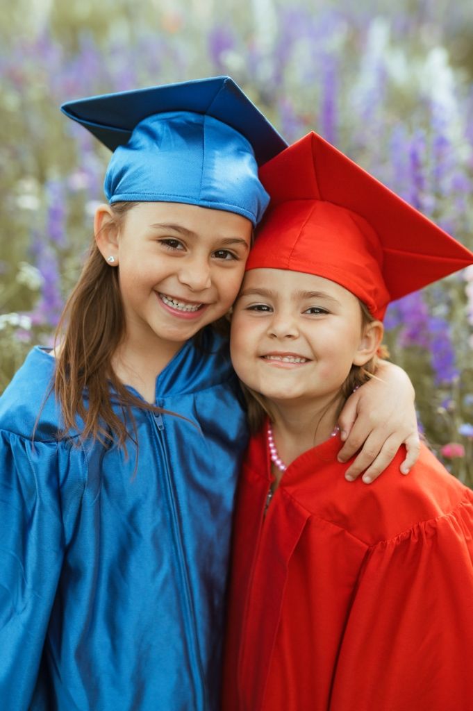 Two graduates in red and blue caps and gowns sharing joyful smiles during a sunny celebration with purple flowers behind them.