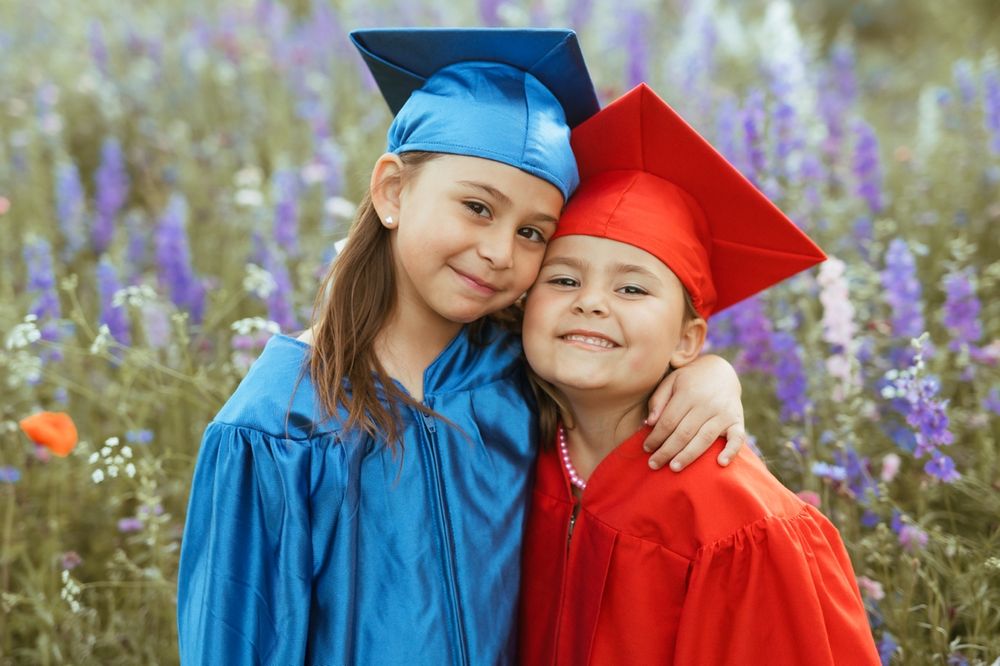 Two young graduates in contrasting blue and red gowns share a joyful moment among blooming wildflowers.