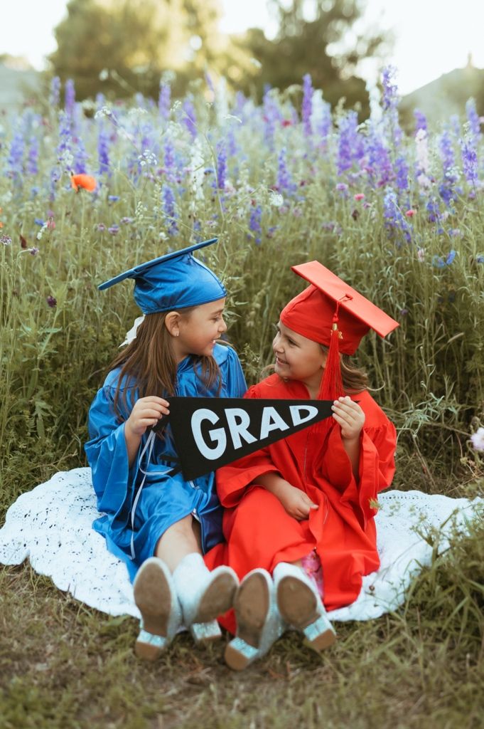Two graduates in red and blue caps and gowns sit together in a wildflower field holding a GRAD sign.