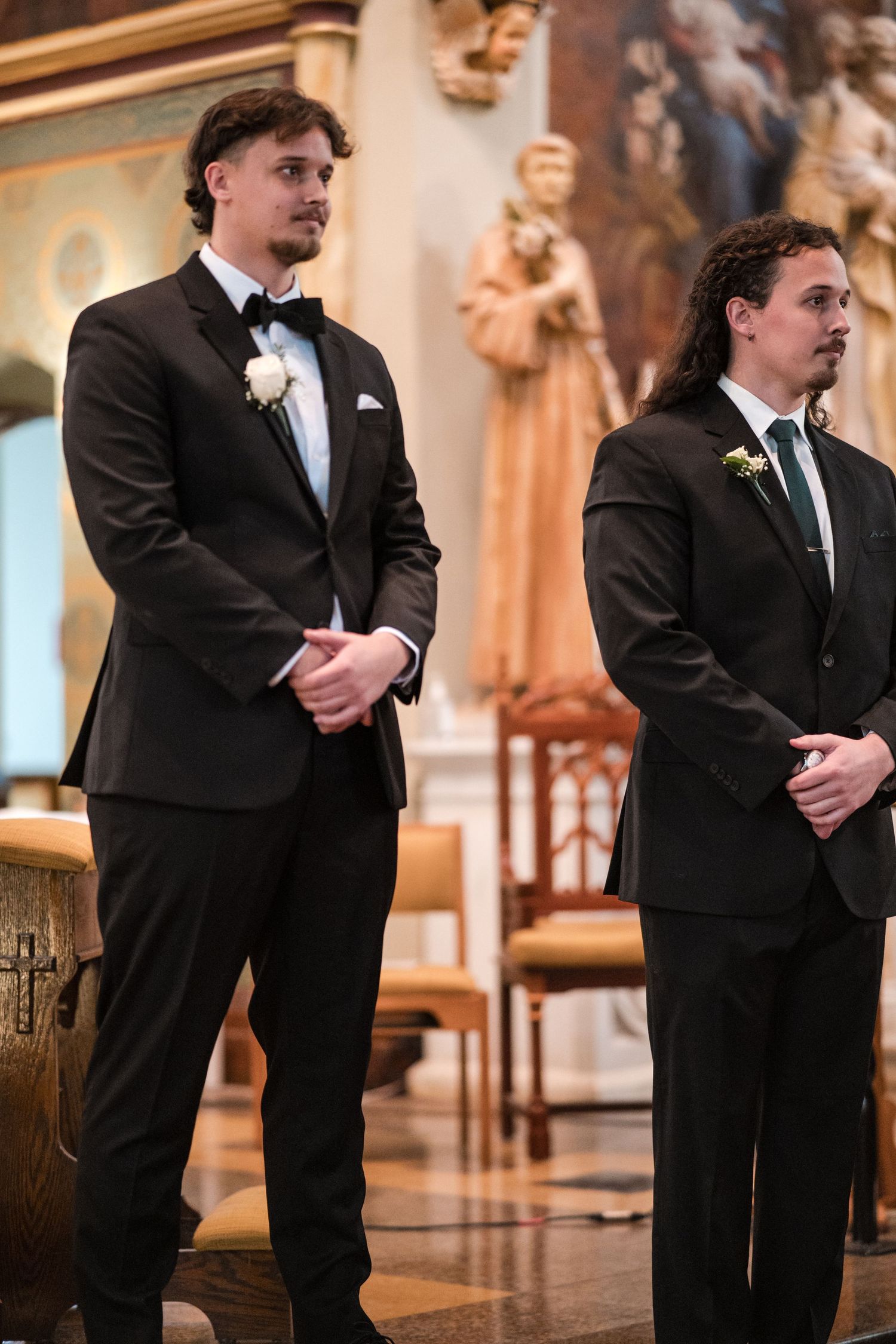Two groomsmen in black tuxedos stand solemnly at the altar in a church during a ceremony.