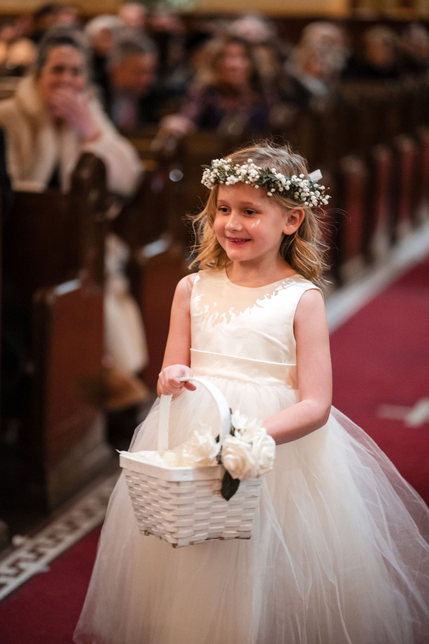 A flower girl in white tulle dress carries a basket down the church aisle during wedding ceremony.