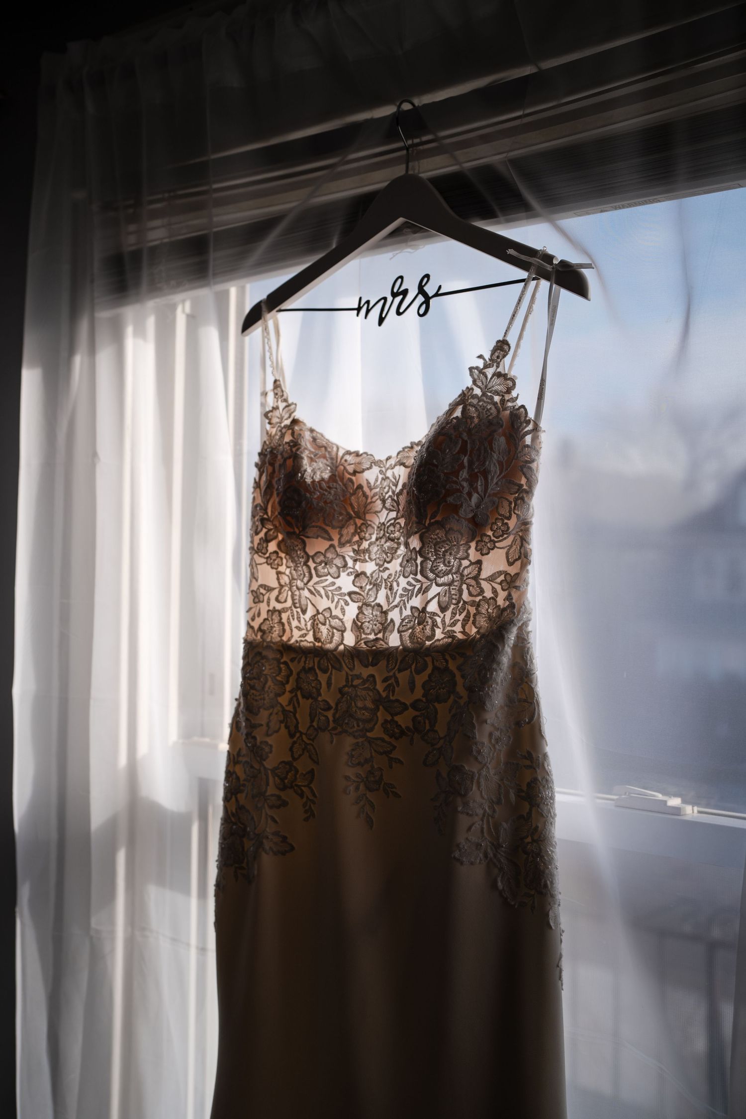Elegant wedding dress with lace detail hanging on custom hanger against backlit window.