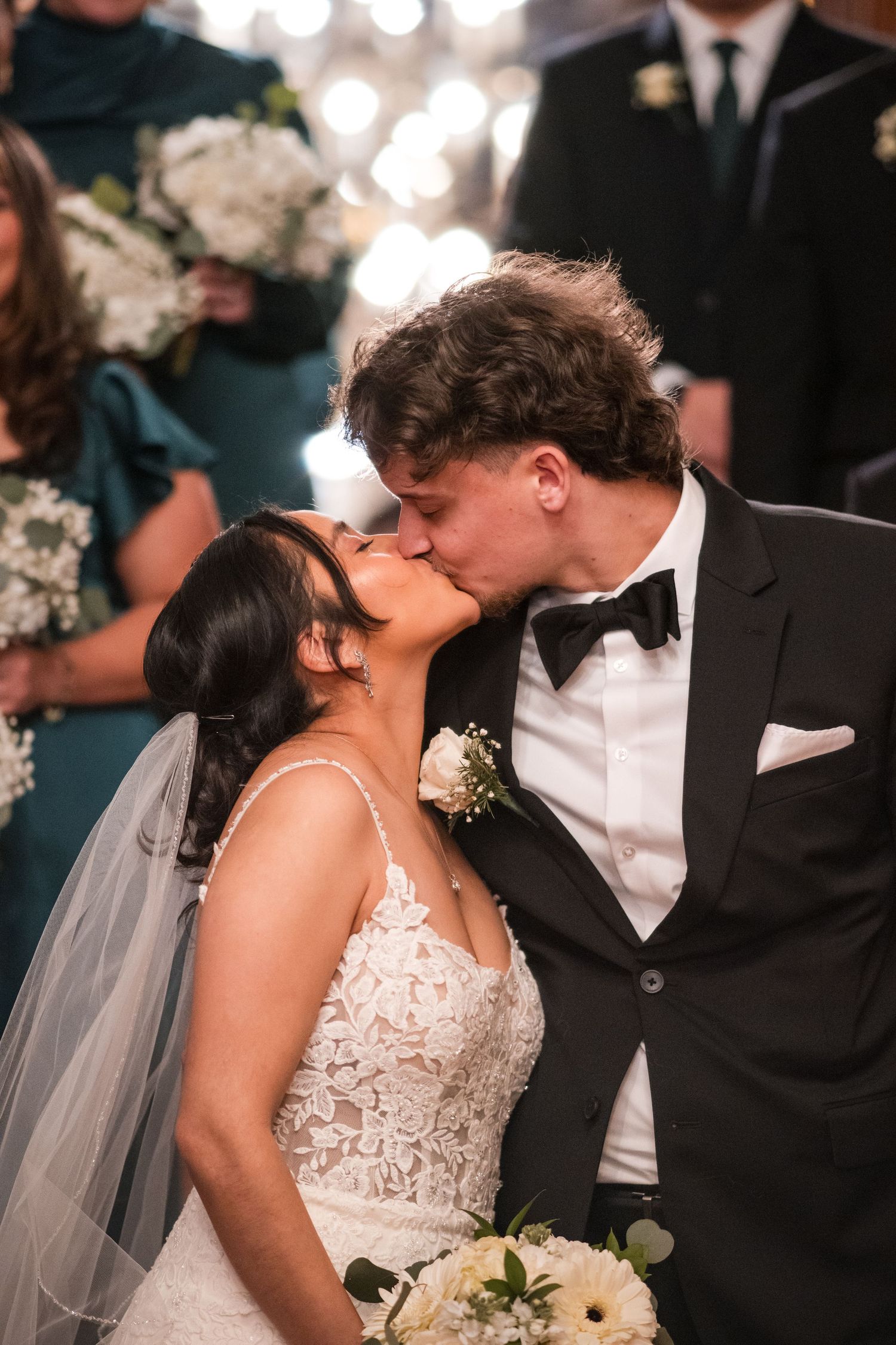 Newlyweds share a romantic kiss during their wedding ceremony with wedding party in background.