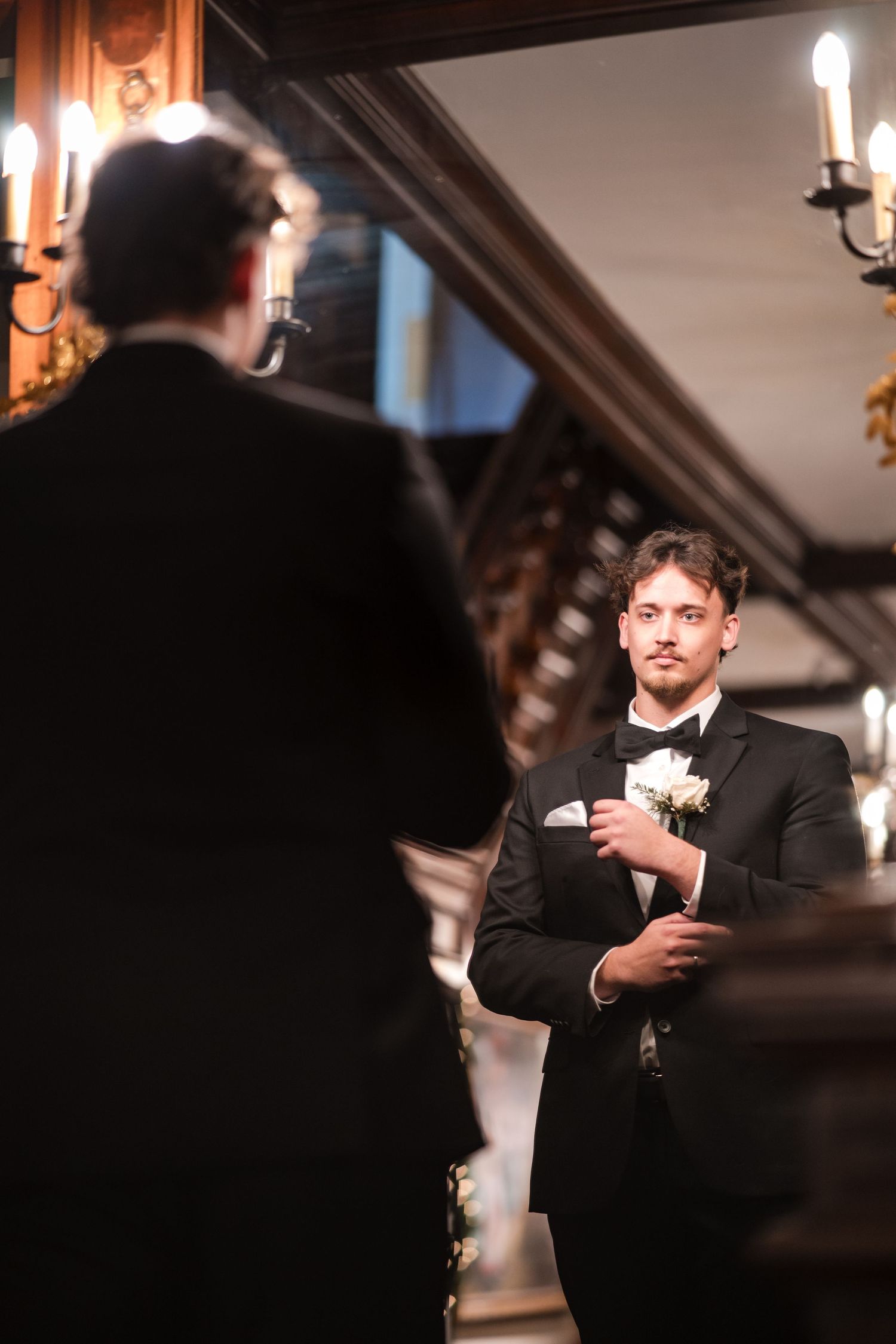 A groom in classic black tuxedo stands thoughtfully in an elegant hallway with chandelier lighting.