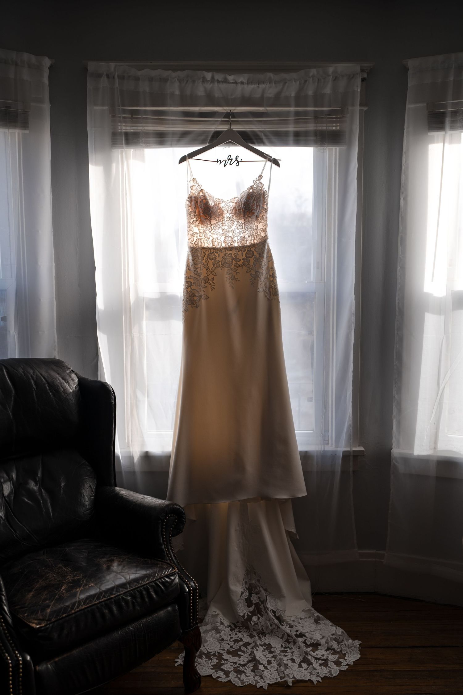 Wedding gown with lace bodice displayed on hanger near leather chair in natural window light.