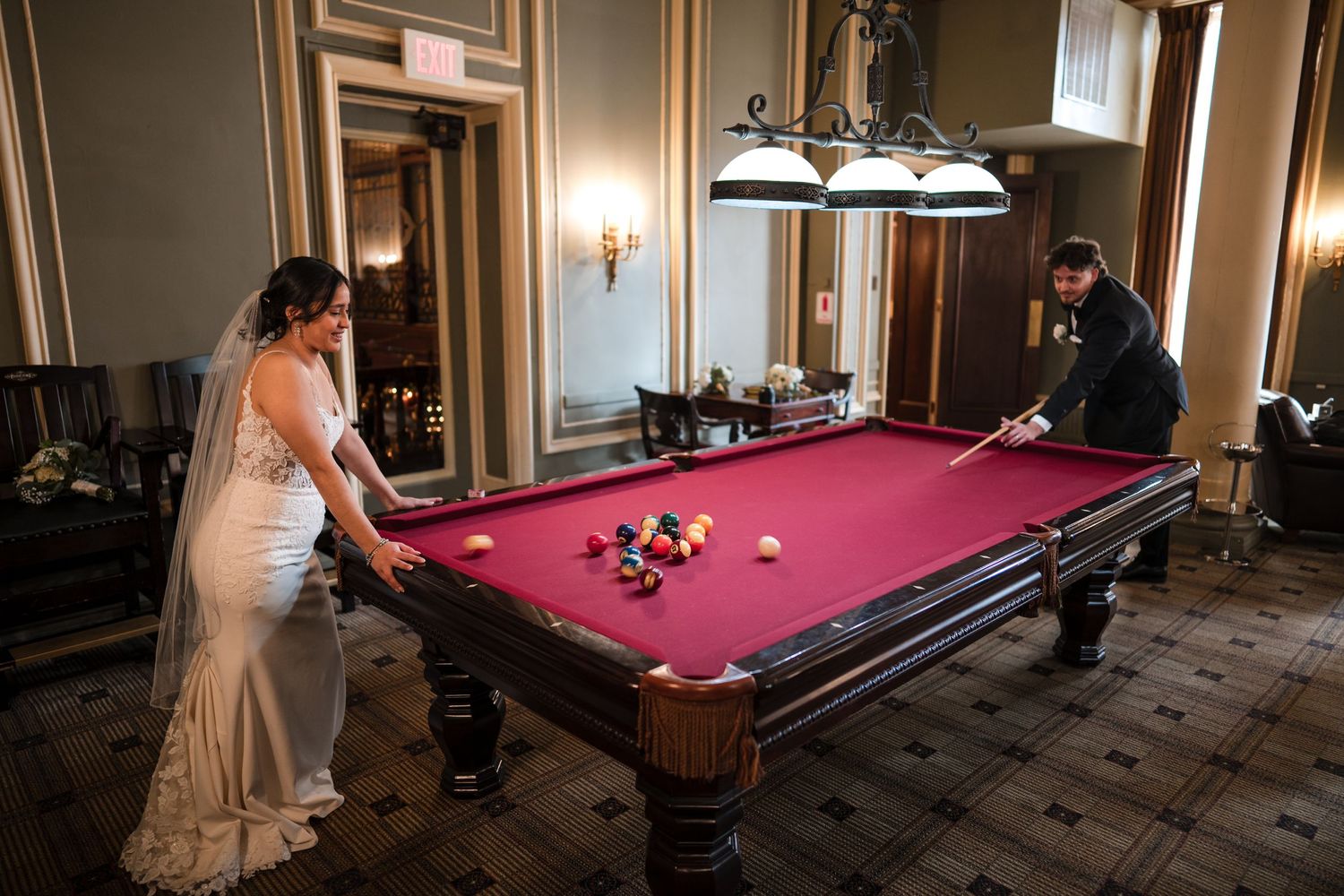Bride in white gown plays pool in an elegant room with dark wood furnishings.