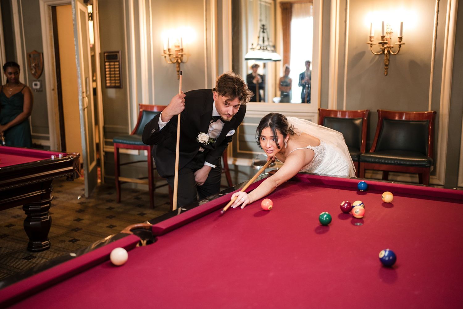 Bride and groom leaning over a red pool table taking shots during their wedding celebration.