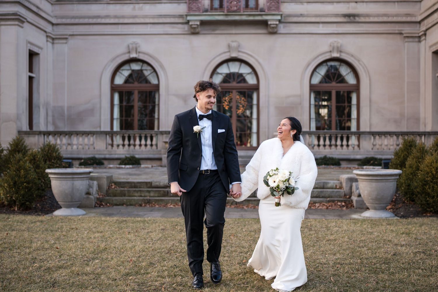 Newlyweds walk together on the lawn of an elegant mansion with arched windows and ornate architecture.