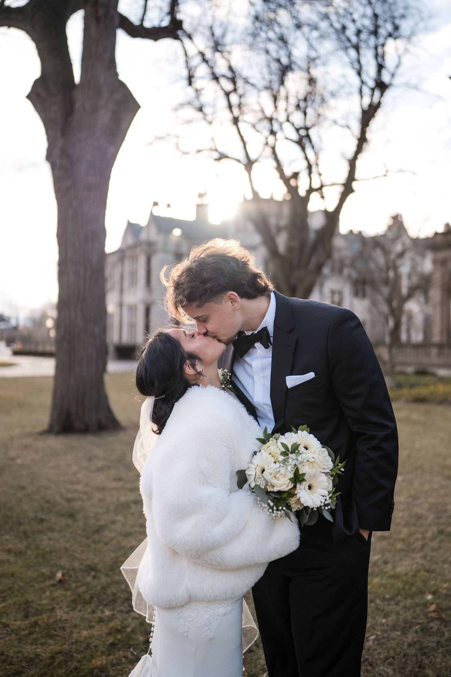 Romantic winter wedding portrait with couple embracing under bare trees at sunset near historic building.