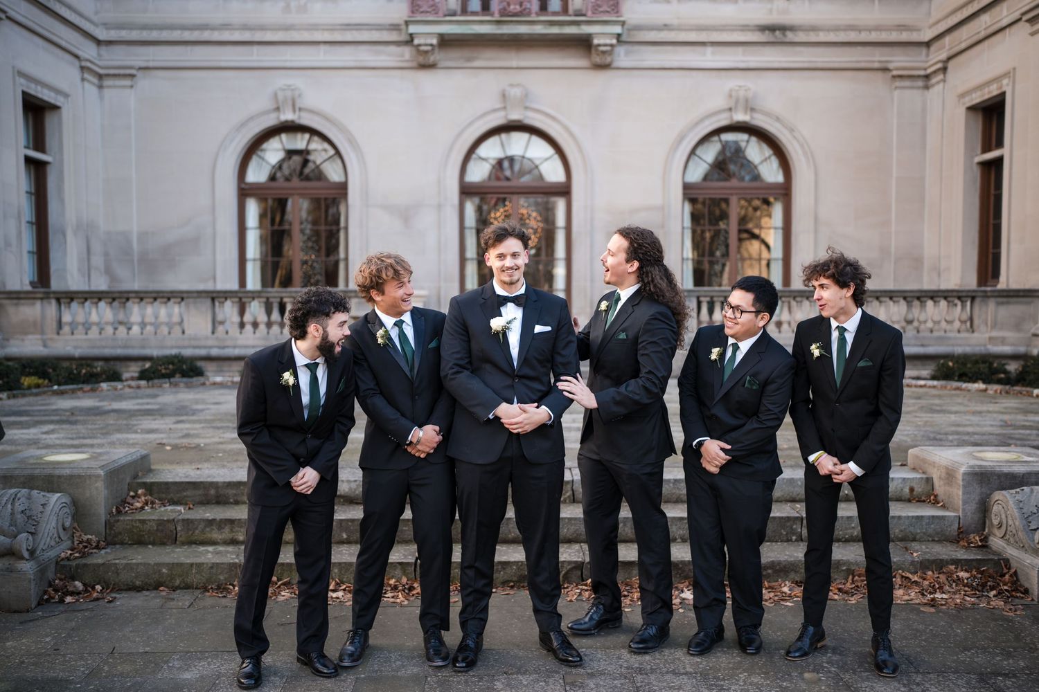 Wedding party in emerald dresses and black suits pose in front of historic building with arched windows.