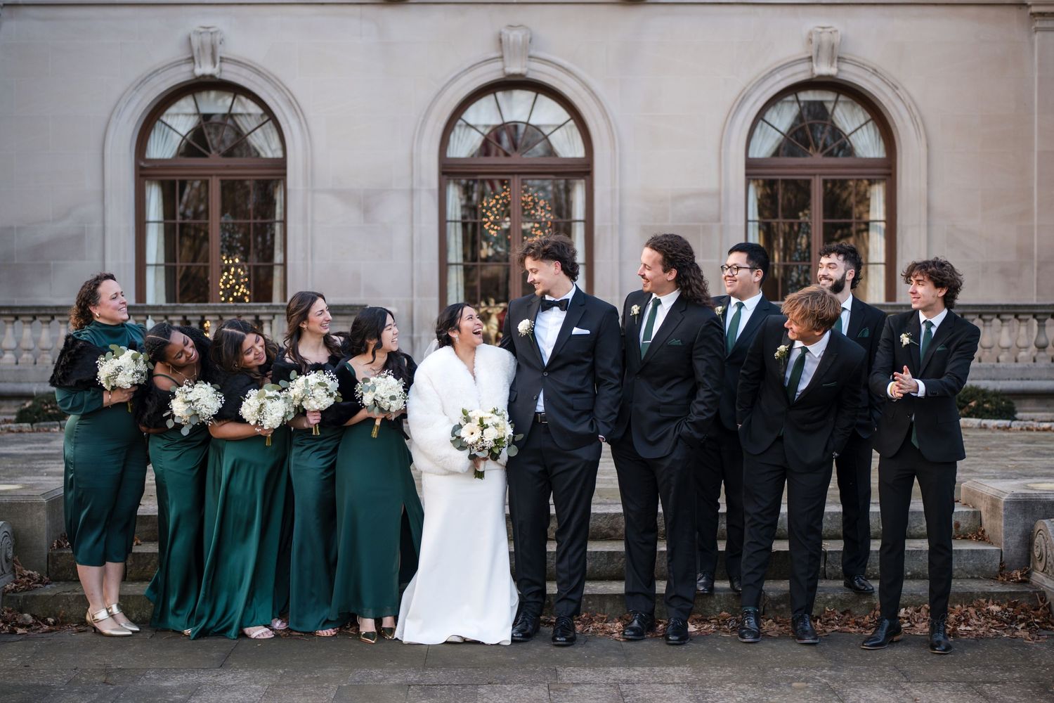 Large wedding party in emerald gowns and dark suits gathered on stone steps of historic venue.
