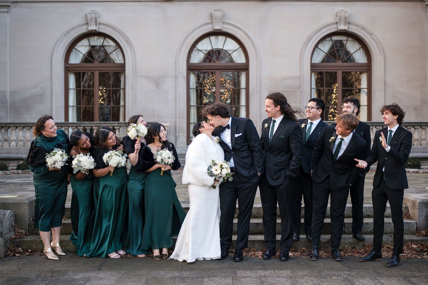 Wedding party dressed in emerald and black share candid moment outside grand historic building.