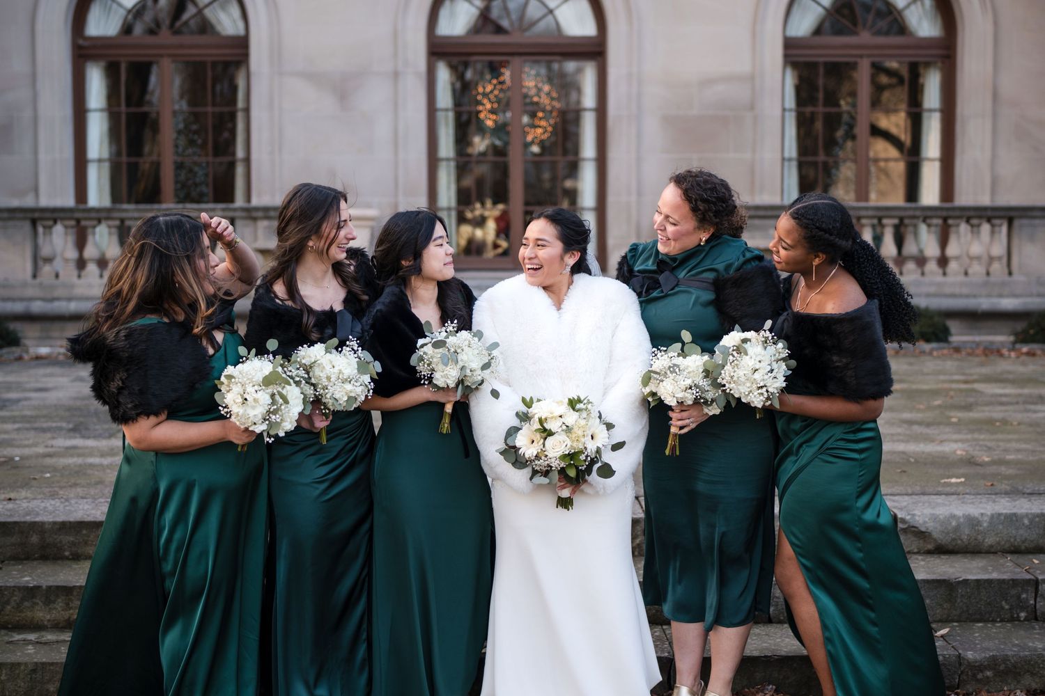 Bridal party in emerald green dresses celebrates joyfully outside historic venue with elegant architecture.