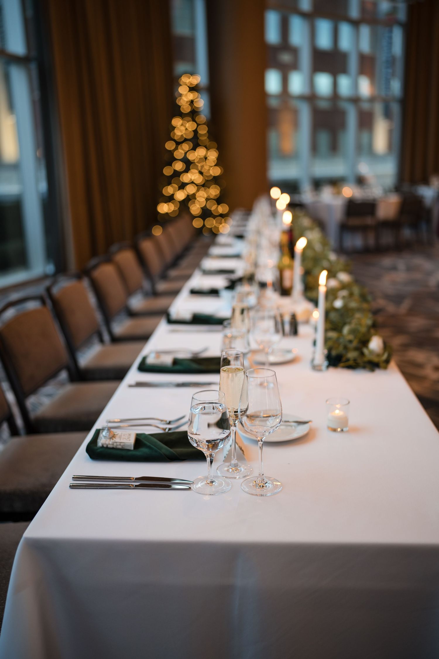 Long banquet table set for wedding reception with white linens and Christmas tree in background.