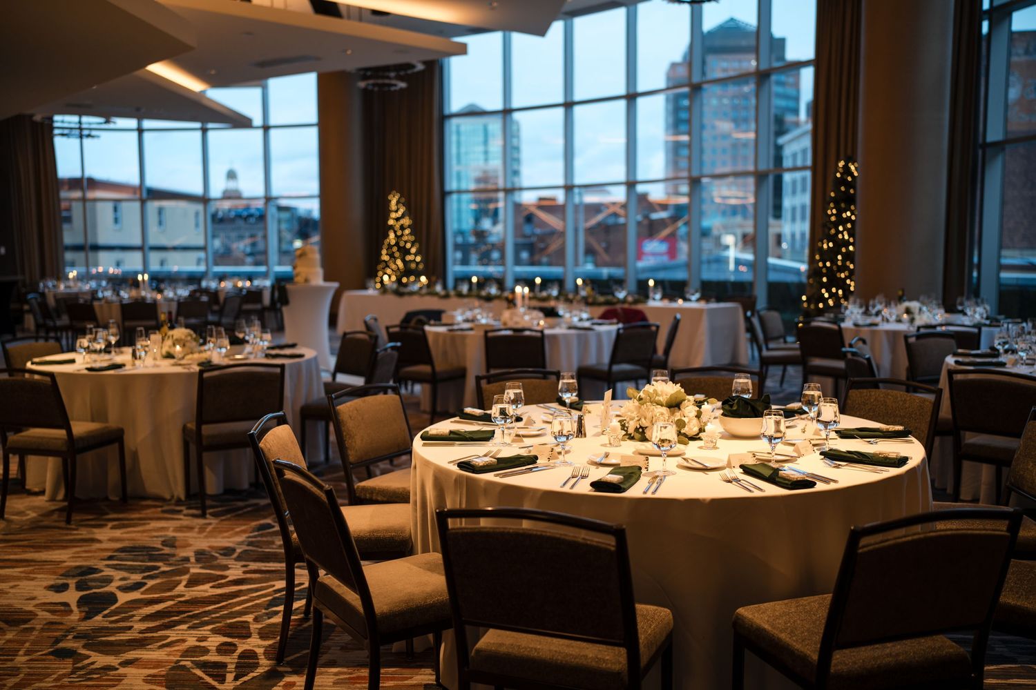 Elegant wedding reception hall with round tables and Christmas tree near floor-to-ceiling windows.