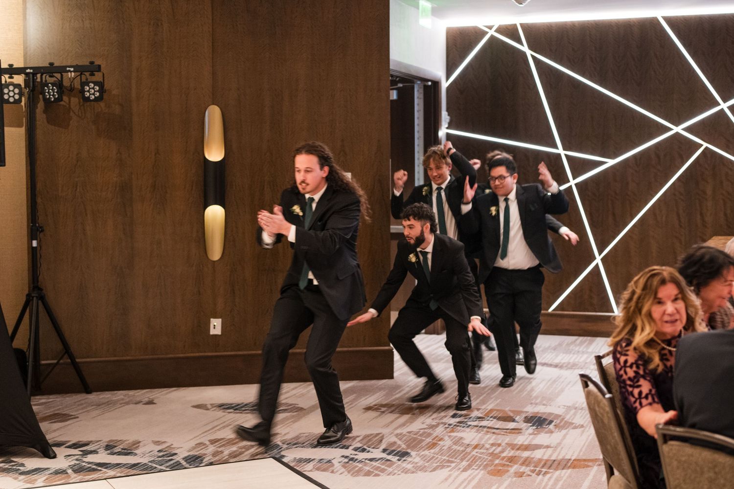 Wedding party dancing energetically in modern venue with geometric light installation.
