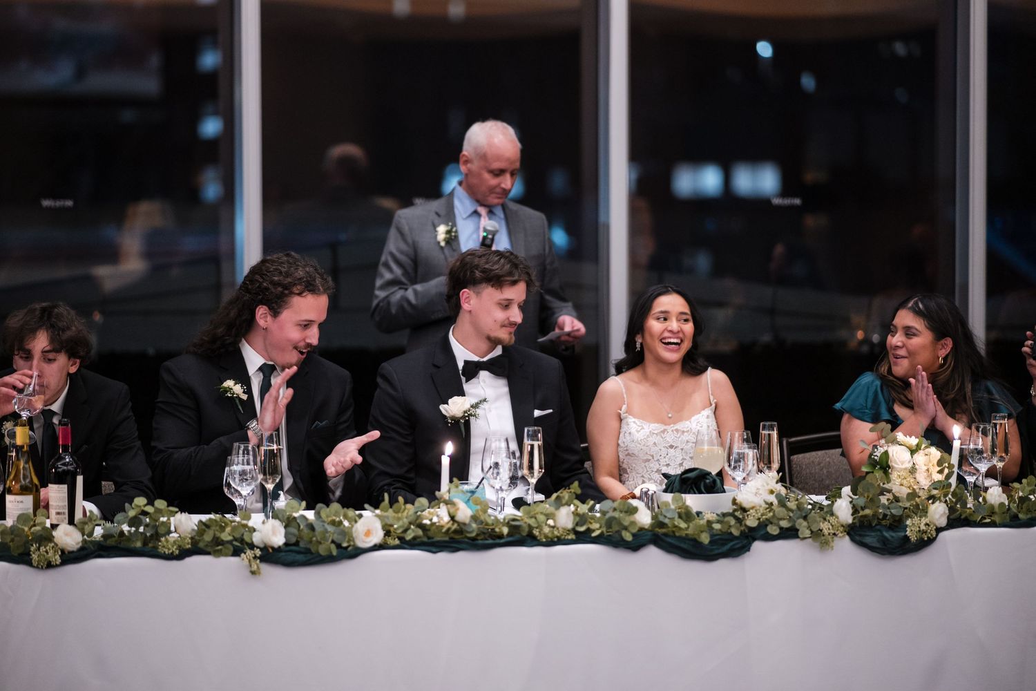 Wedding party seated at head table decorated with floral arrangements at reception.