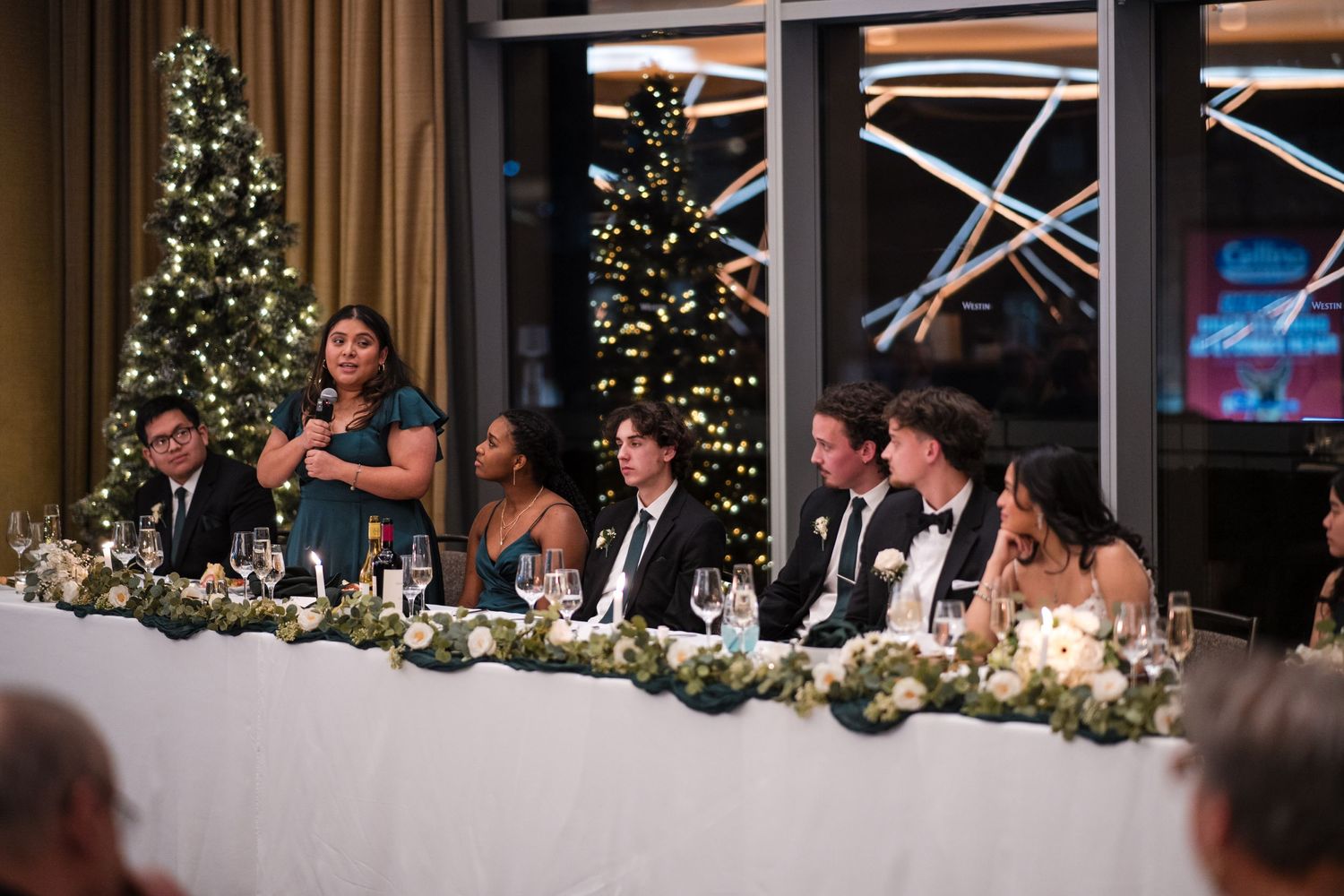 Wedding reception table with guests seated in front of decorated Christmas trees and twinkling lights.