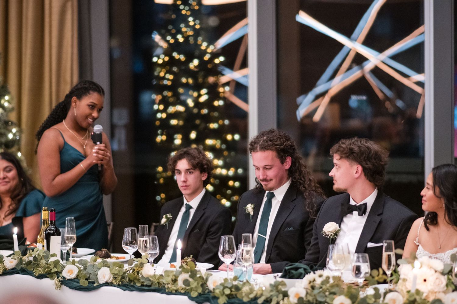 Wedding party enjoys speeches during reception with Christmas tree in background.