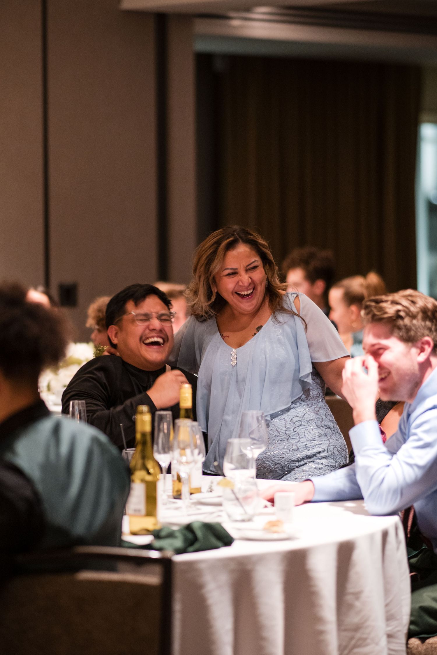 Wedding guests share a moment of genuine laughter during the reception dinner at an elegant indoor venue.