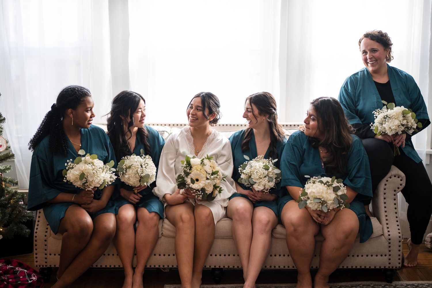 Wedding party in teal dresses sits together holding white bouquets on a light-colored couch.