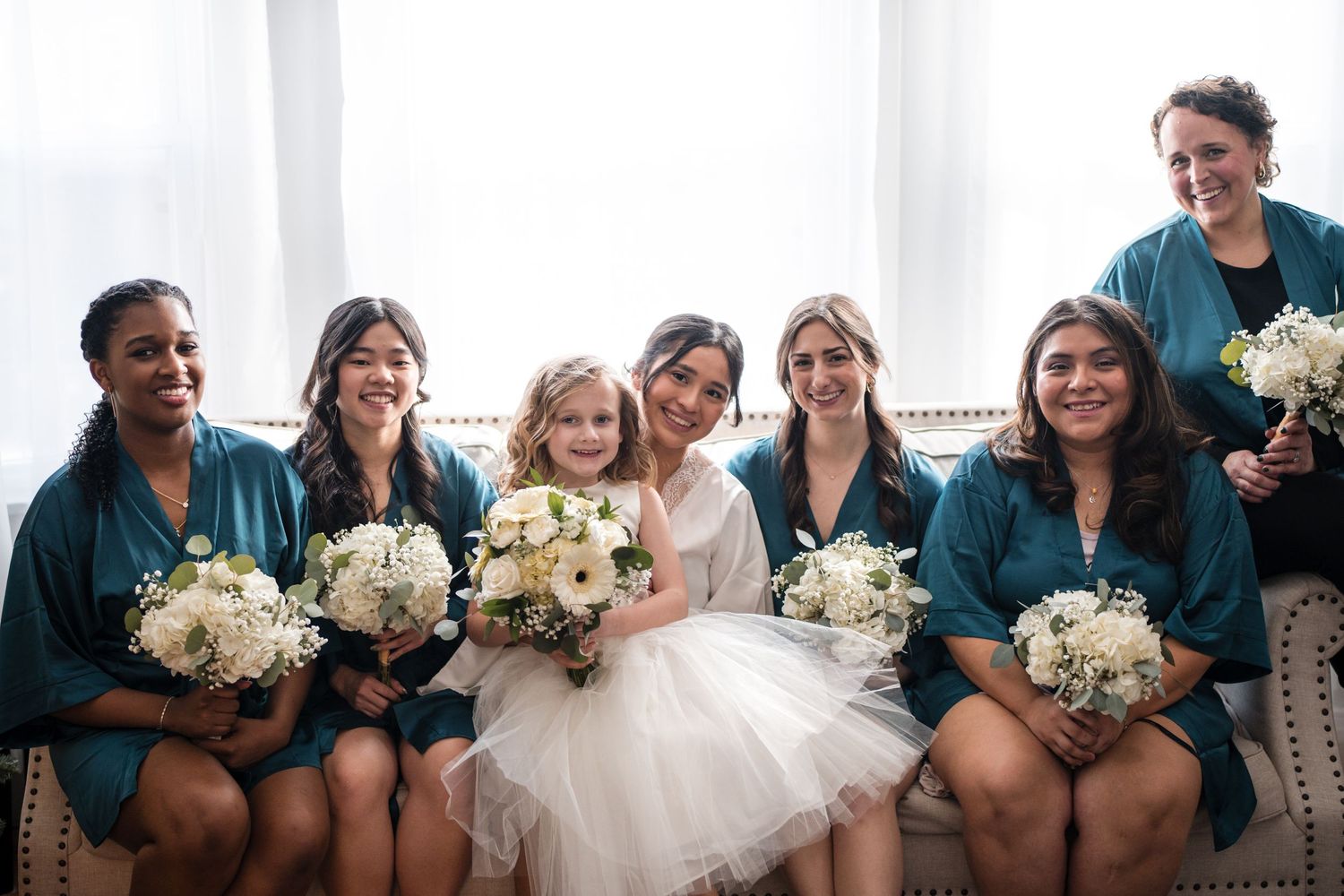 Wedding party poses on couch with bride in white dress and bridesmaids in teal dresses holding bouquets.