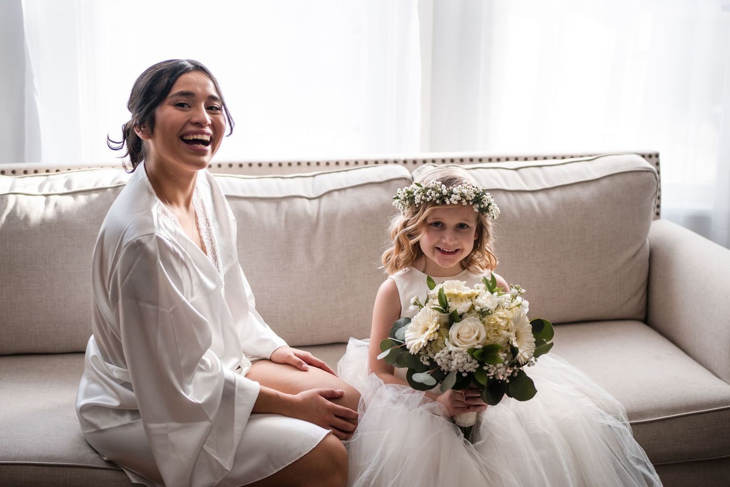 A person in white robe shares a sweet moment with young flower girl on couch.