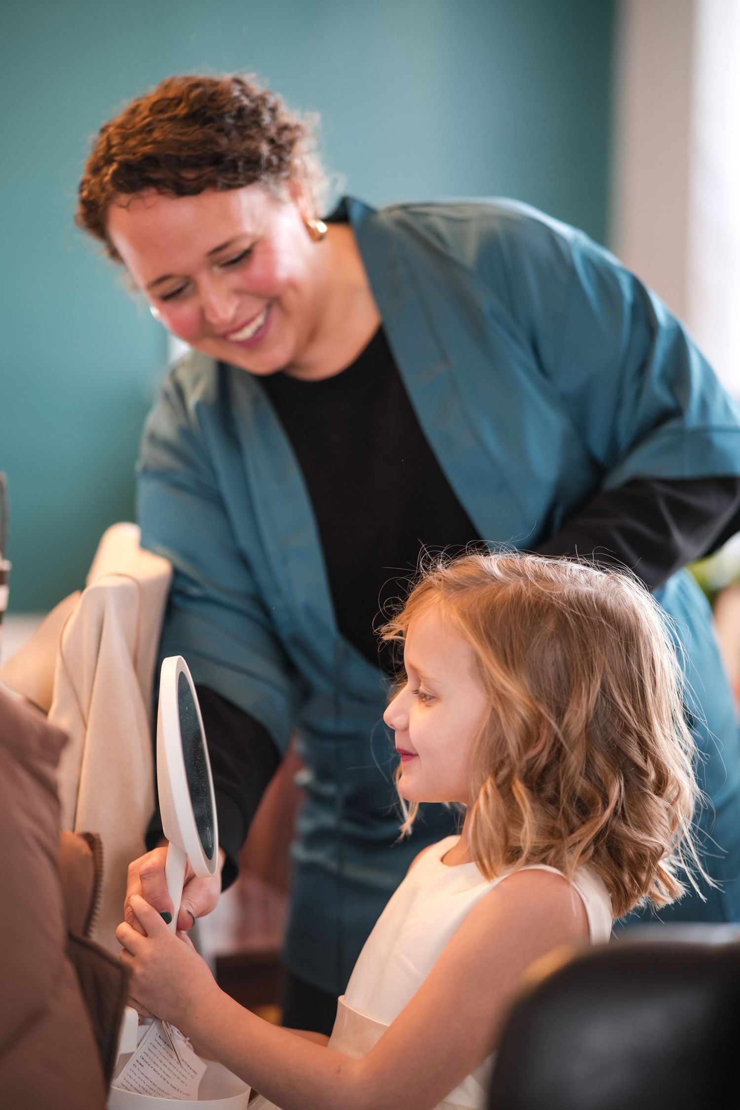 Person in teal jacket watches as young girl looks in mirror during wedding preparations.