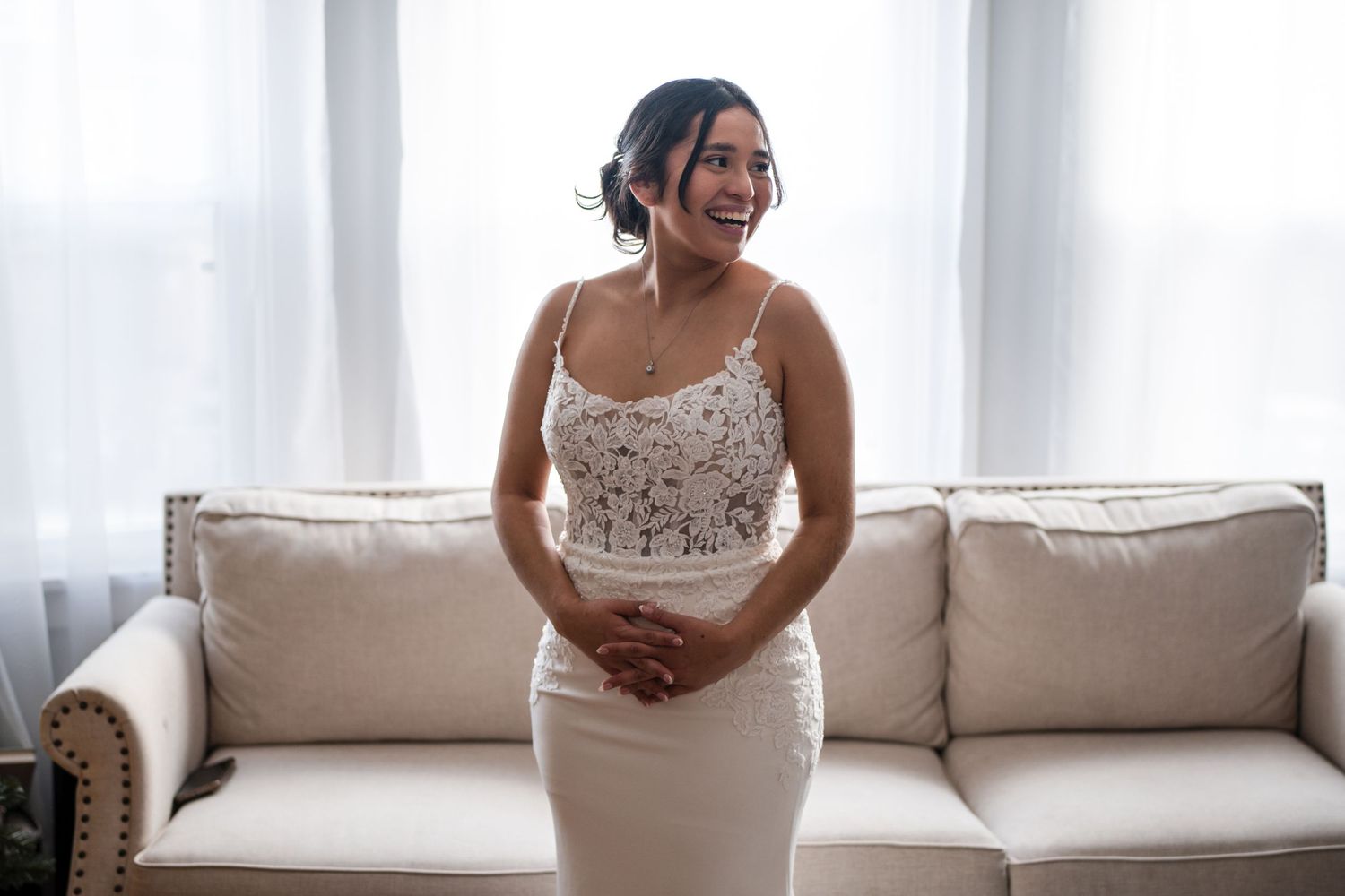 Person in fitted lace wedding gown smiles while standing in front of beige couch.