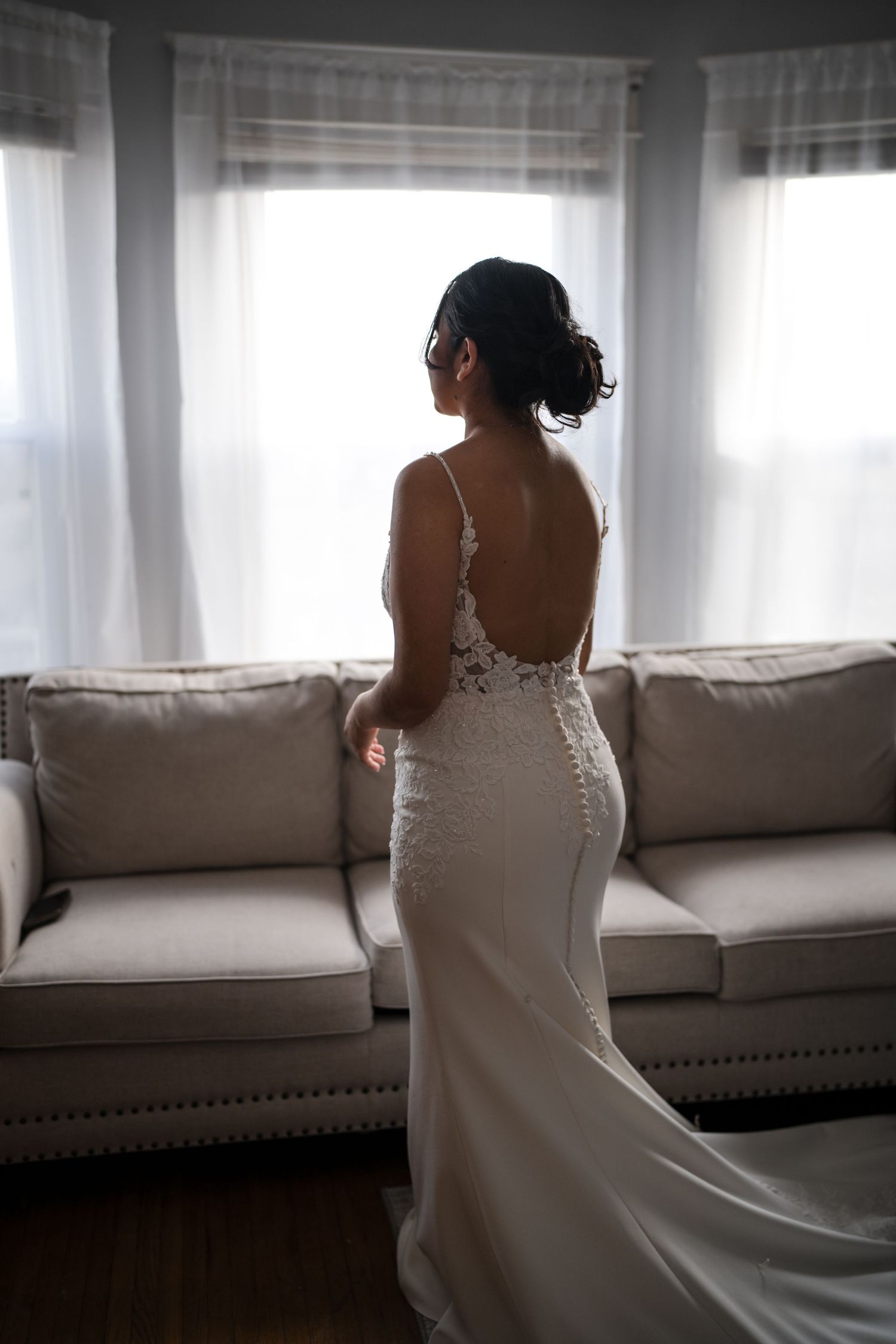 Back view of person in elegant fitted wedding dress with lace details standing by windows.