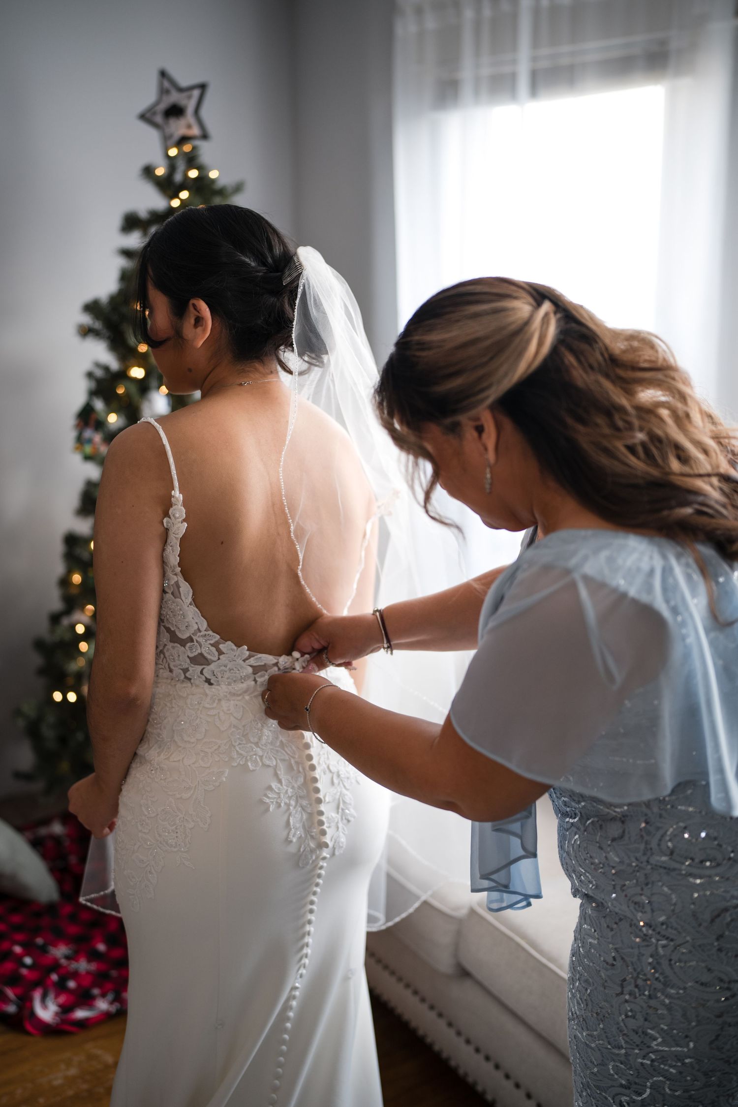 Helper assisting with buttons on back of lace wedding dress with Christmas tree in background.