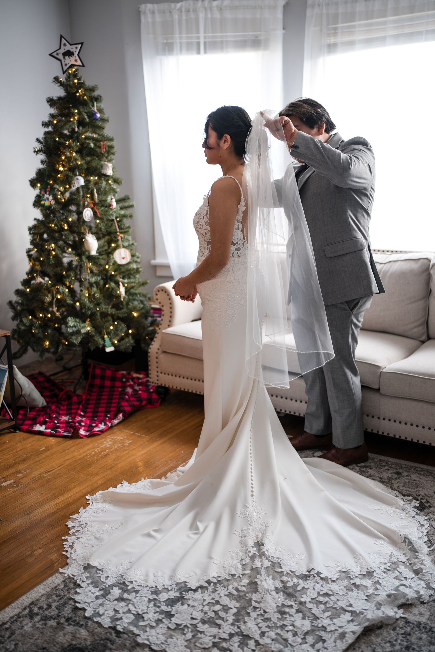 A bride in a white lace gown with a long train stands next to a decorated Christmas tree while getting ready.