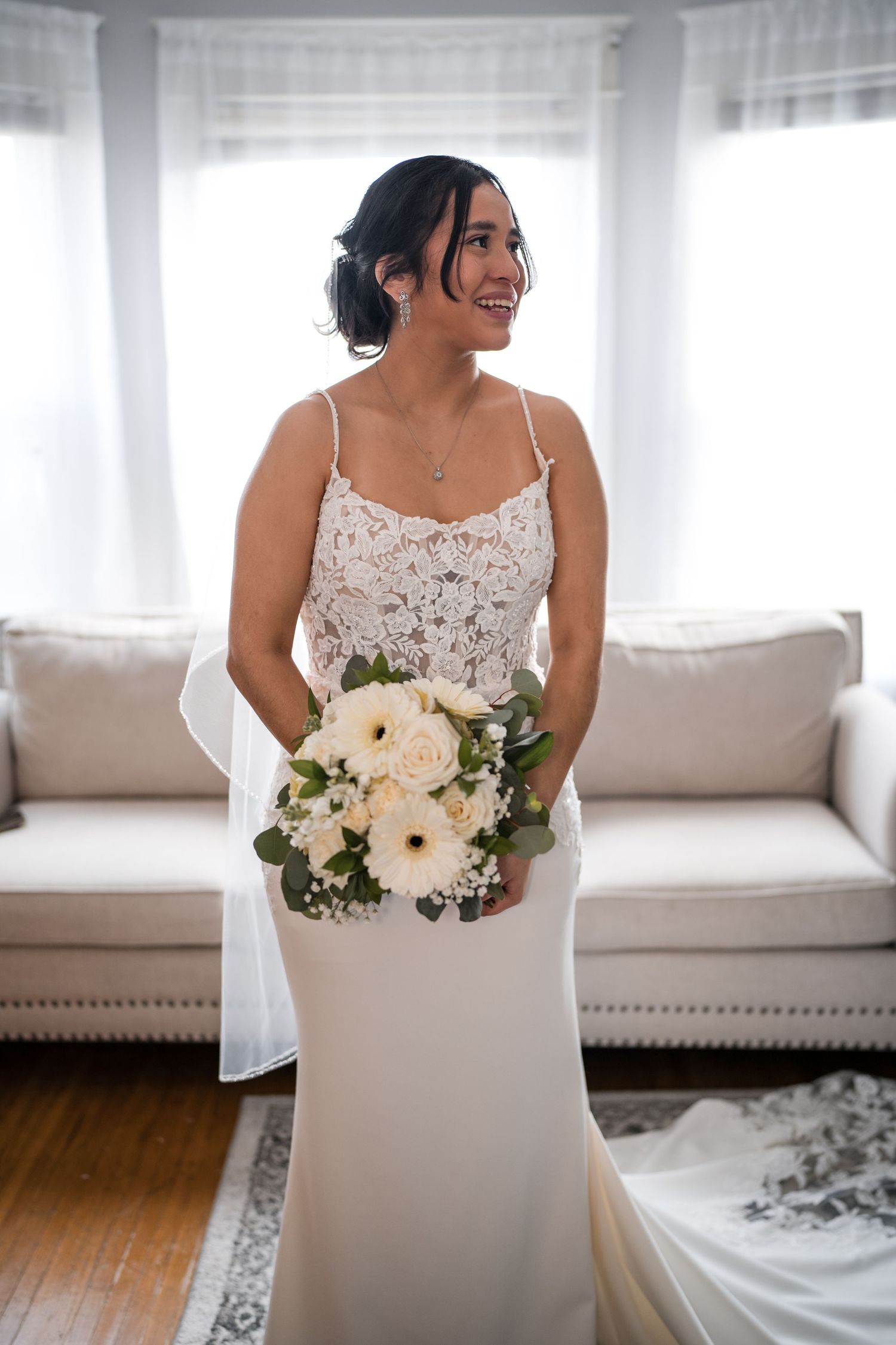 A bride in a fitted white dress holds a bouquet of white flowers while standing in front of sheer curtains.