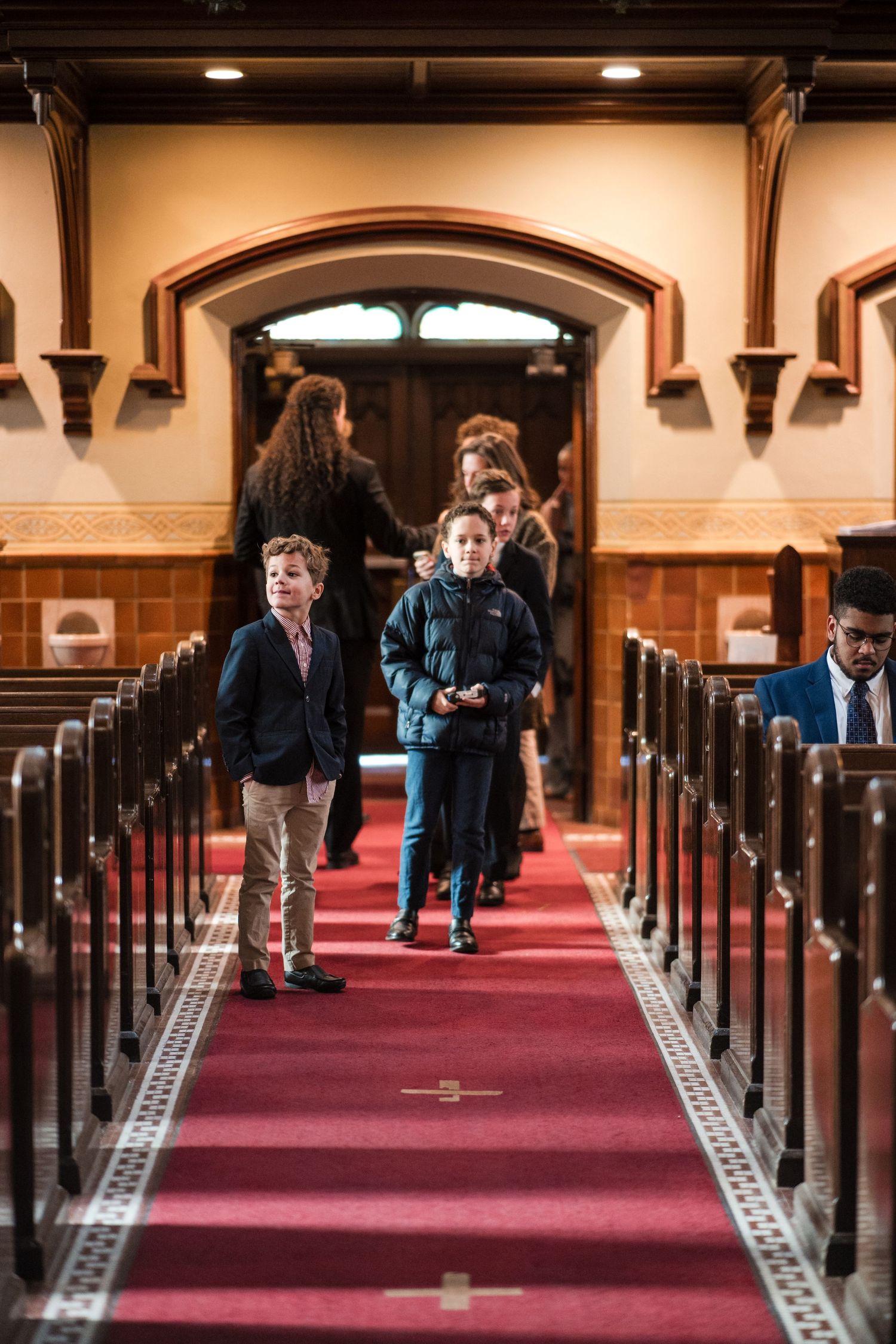 People walk down the red-carpeted center aisle of a church with wooden pews and arched architectural details.