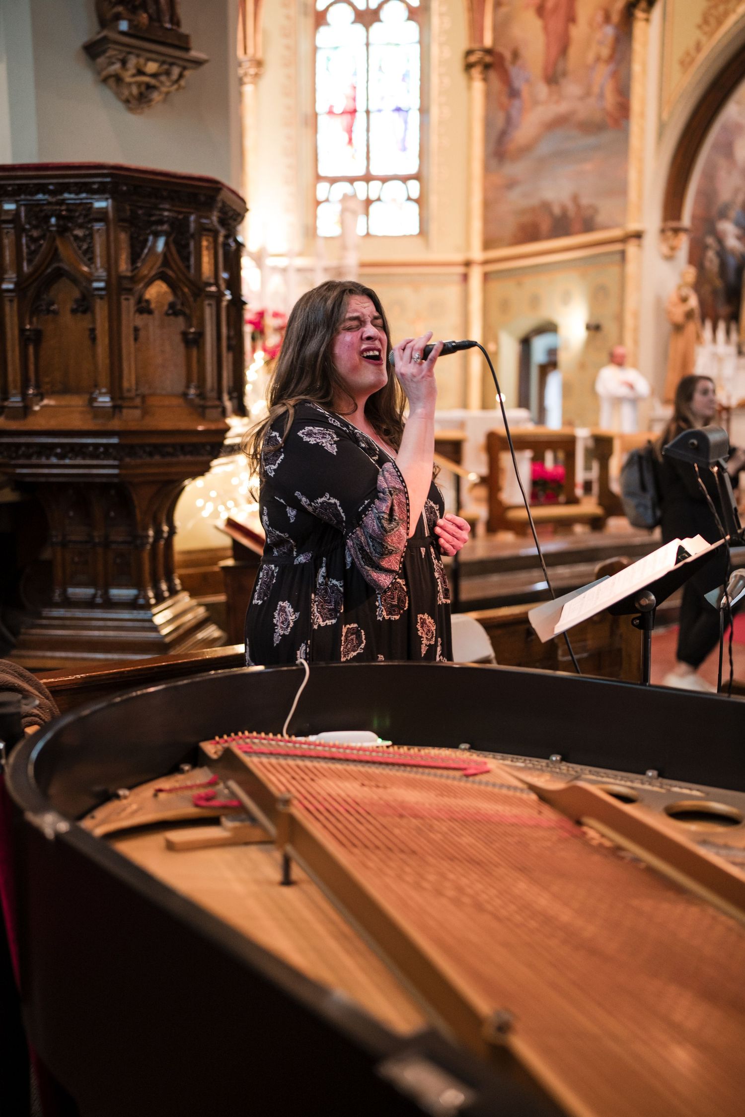 A vocalist performs next to a grand piano in an ornate church setting with stained glass windows.