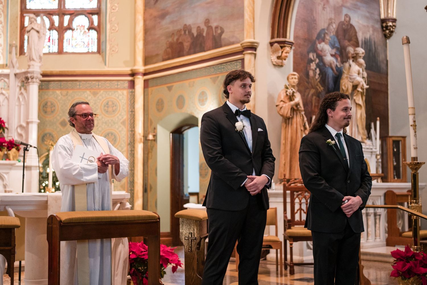 Three people stand at the altar of a church decorated with red poinsettias for a ceremony.