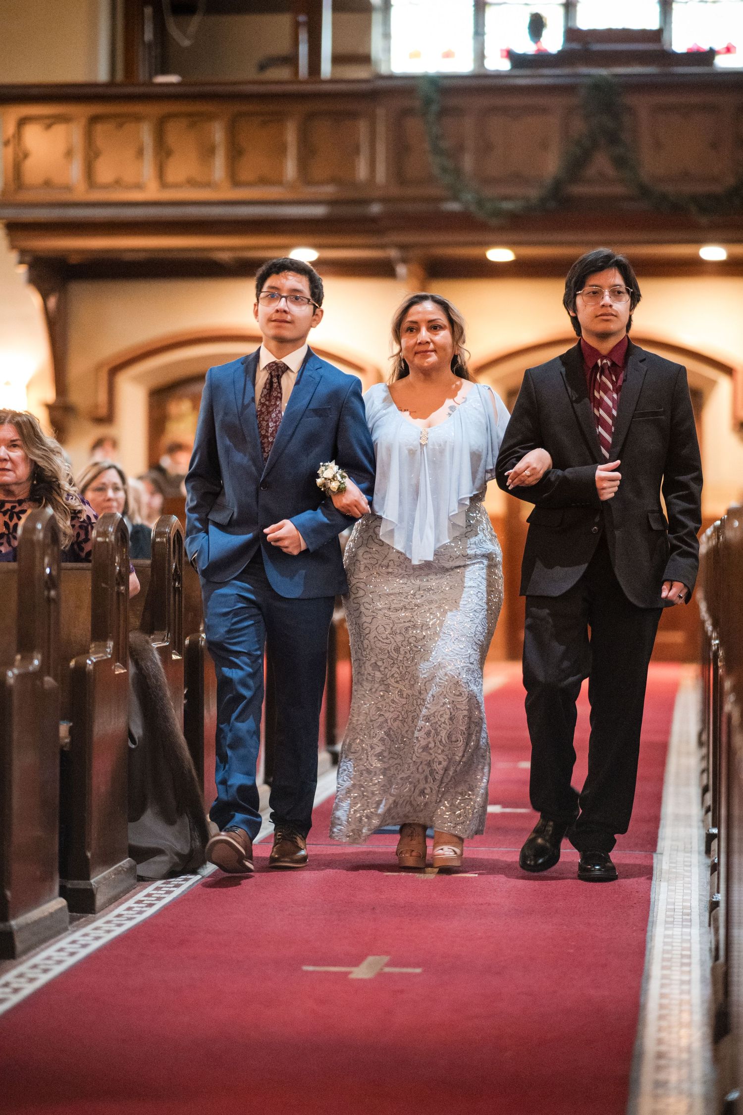 Wedding party members walk down a red-carpeted church aisle lined with wooden pews.